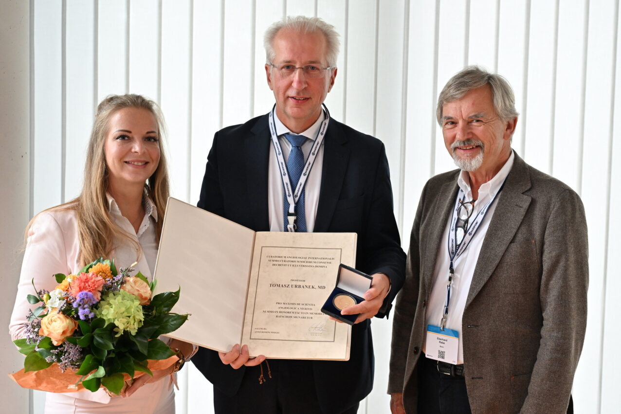 Prof. Dr. Tomasz Urbanek, Gefäßchirurg an der Medizinischen Universität Schlesien in Katowice, steht mit Urkunde und Medaille zwischen zwei Personen. Links hält Michelle Schaub einen Blumenstrauß, rechts steht Prof. Eberhard Rabe. Die Auszeichnung ist die Max-Ratschow-Gedächtnismedaille.